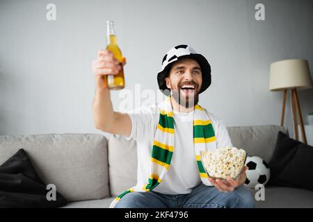 joyeux fan de sport dans un chapeau et un foulard tenant un bol avec pop-corn et une bouteille de bière tout en regardant le championnat, image de stock Banque D'Images