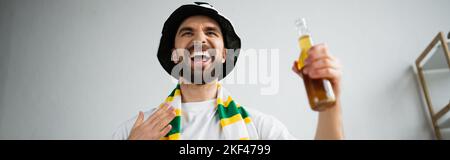 fan de sport émerveillé dans le foulard et le chapeau tenant une bouteille de bière tout en regardant le championnat, bannière, image de stock Banque D'Images