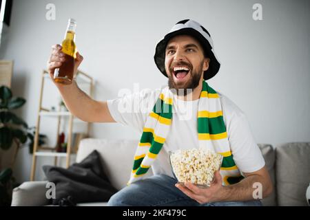 fan de sport enthousiaste dans un chapeau et un foulard tenant un bol avec pop-corn et une bouteille de bière tout en regardant le championnat, image de stock Banque D'Images