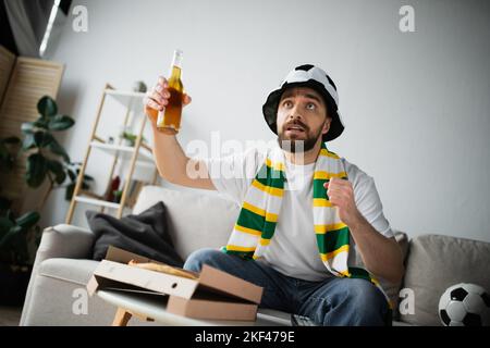 homme tensionné dans un chapeau de fan sportif et un foulard tenant une bouteille de bière tout en regardant le championnat, image de stock Banque D'Images