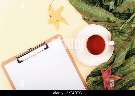 Bureau avec presse-papiers vierge, une tasse de thé, des feuilles jaunes et une écharpe verte sur fond beige clair. Vue de dessus, plan d'installation, maquette. Banque D'Images