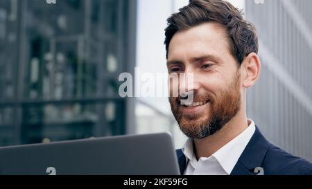 Gros plan souriant homme d'affaires réussi travaillant sur un ordinateur portable en plein air joyeux homme barbu Manager indépendant communiquant à distance avec le client Banque D'Images