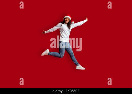 Jolie femme noire émotive portant un chapeau de père Noël sautant sur le rouge Banque D'Images