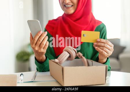 Femme musulmane moderne et mature tenant une carte de crédit et un téléphone portable, déballant une boîte en carton avec un nouveau casque Banque D'Images