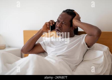 Frustré Black Man parlant sur le téléphone portable tout en étant assis dans le lit à la maison Banque D'Images