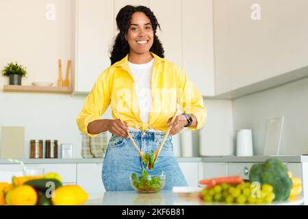 Préparer des repas sains. Une femme américaine africaine positive qui prépare une salade fraîche saine et souriait à l'appareil photo dans la cuisine Banque D'Images