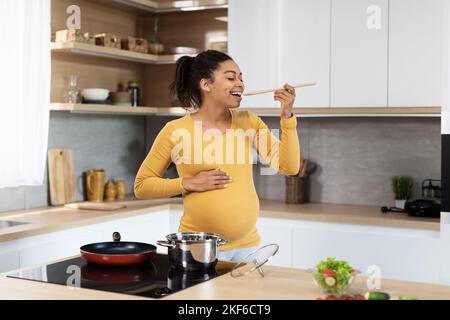 Une jeune femme enceinte noire souriante au ventre géant prépare le dîner, goûte la soupe dans un intérieur minimaliste de la cuisine Banque D'Images