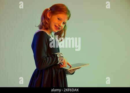 Portrait d'une jolie petite fille à tête rouge portant une robe de fête à l'aide d'une tablette isolée sur un fond clair dans une lumière néon. Émotions des enfants, visage Banque D'Images