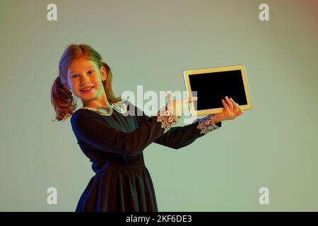 Portrait d'une jolie petite fille à tête rouge portant une robe de fête à l'aide d'une tablette isolée sur un fond clair dans une lumière néon. Émotions des enfants, visage Banque D'Images