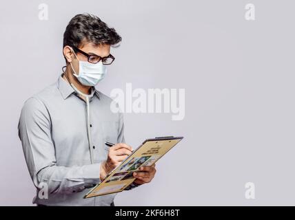 Cliché isolé d'un médecin d'Asie du Sud portant un masque facial et écrivant sur un écritoire sur fond gris Banque D'Images
