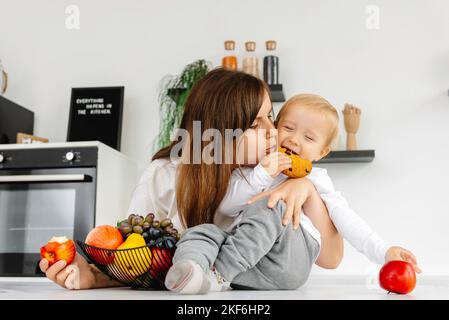 Bonne famille maman embrassant bébé et mangeant des pommes et des aliments sains. Repas du Réducétaire en savourant des aliments végétaux Banque D'Images