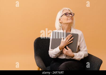 Belle femme sénior attentionnés en costume d'affaires assise sur un fauteuil avec un ordinateur portable et pensant sur un fond marron isolé Banque D'Images