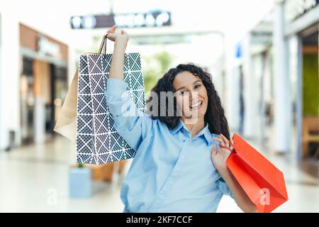 Bonne jeune femme hispanique attrayante tenant des sacs de shopping colorés dans ses mains dans le centre commercial, dansant, réjouissant, levant les sacs. En regardant l'appareil photo, en souriant. Banque D'Images