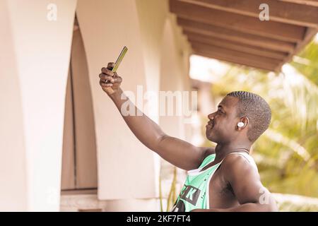 Vue latérale d'un touriste noir qui écoute de la musique avec de véritables écouteurs sans fil et qui prend son selfie sur la terrasse de l'hôtel le jour de l'été sur la station Banque D'Images