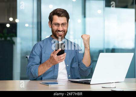 Joyeux jeune homme beau indépendant jouant des jeux en ligne sur le téléphone au travail. Lève la main, montre oui, gagné. Assis dans le bureau au bureau. Banque D'Images