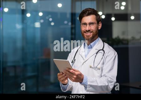 Portrait d'un jeune homme médecin debout sur un manteau blanc et avec un stéthoscope dans un bureau dans un hôpital, tenant une tablette dans ses mains, regardant la caméra, souriant. Banque D'Images