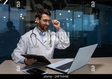Un jeune médecin de sexe masculin parle à un patient en utilisant un appel vidéo depuis un ordinateur portable. Il est assis à une table dans un bureau d'un hôpital. Il tient un dossier avec des documents, ajuste ses lunettes, parle, sourit. Banque D'Images