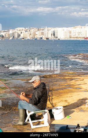 Sliema, Malte - 12 novembre 2022: Pêcheur maltais assis sur une chaise sur les rochers au bord de la mer tout en pêchant avec un bâton de pêche le matin ensoleillé Banque D'Images