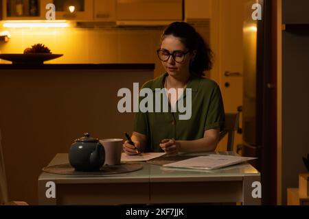 Freelance et travail à distance. Jolie jeune femme caucasienne portant des lunettes est assise à table et comptable. Soirée à la maison. Lady fait ses devoirs. Co Banque D'Images