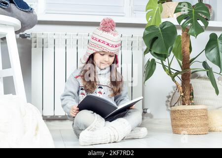 Intéressé, sérieuse petite fille mignon en laine chapeau et vêtements chauds avec bonhomme de neige livre de lecture à côté du radiateur de chauffage dans le salon blanc. Confortable et Banque D'Images