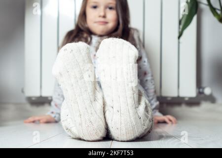 Gros plan, concentrez-vous sur les pieds de petite fille souriante et calme dans des chaussettes blanches en laine, montrant à l'appareil photo, assis près du radiateur de chauffage à la maison. Préchauffage après Banque D'Images