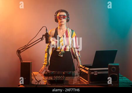 Jeune musicien souriant portant des lunettes de soleil fraîches à la fête sutdio, mélangeant de la musique techno aux platines et utilisant des vinyles. Artiste en train de faire des performances à la boîte de nuit avec équipement audio et mixeur. Banque D'Images