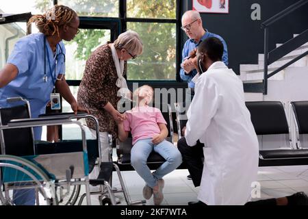 La petite fille s'évanouisse et s'effondre sur la chaise dans la salle d'attente de l'hôpital pendant la consultation médicale, les grands-parents effrayés essayant d'aider. Médecin praticien consultant l'enfant essayant de la réveiller Banque D'Images