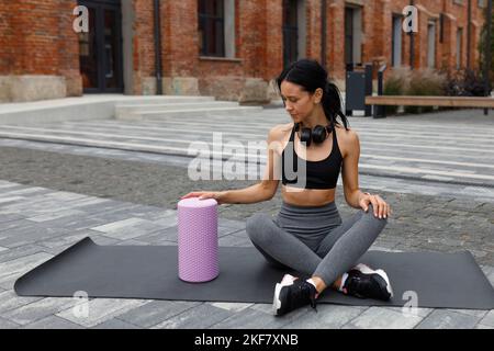 Femme de fitness dans les vêtements de sport assis sur le tapis de yoga et prend le rouleau de massage pendant l'exercice à l'extérieur Banque D'Images