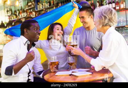 Des amis heureux de différents âges boivent de la bière et célèbrent la victoire de l'équipe ukrainienne dans le bar de nuit Banque D'Images