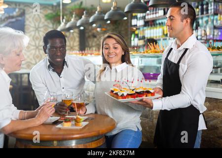 Un serveur latin positif en tablier pointe la main sur l'assiette avec des pinchos au milieu de la barre Banque D'Images