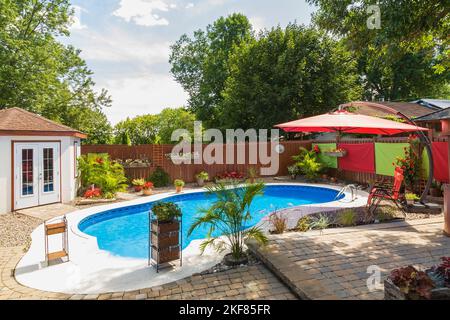 Piscine ovale irrégulière bordée de pavés de couleur Havane et entourée de plantes tropicales et de fleurs dans l'arrière-cour. Banque D'Images