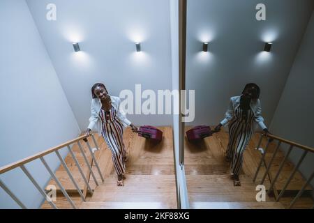 Une photo en grand angle d'une jeune charmante jeune femme noire qui monte les escaliers avec ses valises à roulettes roses ; une jeune femme africaine joyeuse est à la mode Banque D'Images