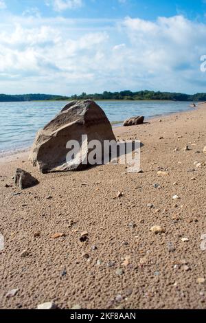 Une grande roche sur la rive sablonneuse d'un lac. L'eau, les arbres et le ciel bleu sont en arrière-plan. Banque D'Images