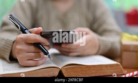 Gros plan et court, décontracté jeune femme asiatique dans un pull confortable utilisant son smartphone pour rechercher des informations en ligne tout en lisant un livre. Banque D'Images