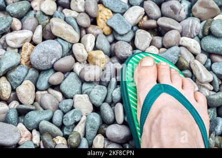 Pied touristique dans des tongs vert clair se dresse sur la plage de galets près de la mer. Un homme se tient sur la côte humide pour se détendre en gros plan Banque D'Images