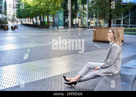 Femme d'affaires rêveuse en costume, assise dans une rue vide près des immeubles de bureaux, en regardant loin Banque D'Images