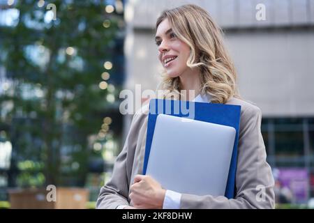 Portrait d'une jeune femme d'affaires, directeur de bureau marchant dans la rue avec un ordinateur portable et un dossier de travail, souriant Banque D'Images