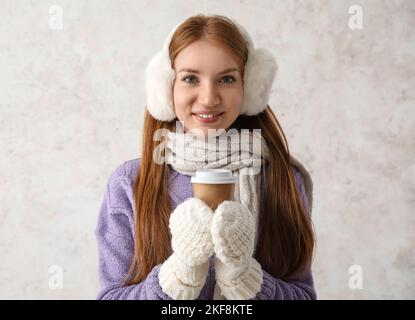 Jeune femme à tête rouge dans des couvre-oreilles chauds avec une tasse de café sur fond clair Banque D'Images