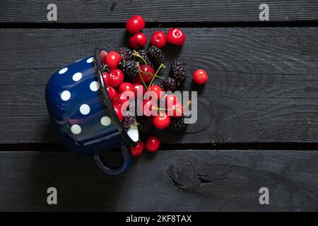 cerises et mûres dans une tasse sur une table en bois noir parsemée, fond de fruits, jus de fruits Banque D'Images