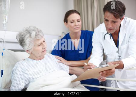 Heres une mise à jour sur votre état... Un médecin et son infirmière debout sur le lit de leurs patients tout en lui montrant le rapport médical. Banque D'Images