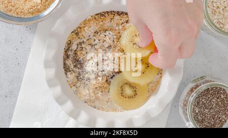 Femme à la main plaçant des tranches de kiwi doré sur une assiette avec des flocons d'avoine instantanés, à plat Banque D'Images
