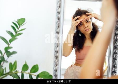 Inquiet afrique jeune fille vérifiant problème de peau de visage regarder dans le miroir. La jeune femme touche le visage squah pop zit bouton sur le front enfoncé par la tête noire Banque D'Images