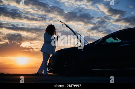 Silhouette de femme à la recherche d'un service d'assistance routière pour résoudre les problèmes de voiture électrique sous le capot en utilisant la technologie de smartphone pendant le coucher du soleil, la femme pilote ont l'assistance en ligne Banque D'Images