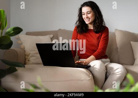 Jeune femme heureuse utilisant un ordinateur portable assis sur un canapé à la maison entouré de plantes Banque D'Images