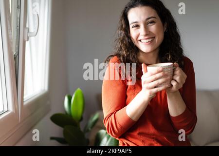 Jeune belle femme riant tout en ayant une boisson chaude près de la fenêtre à la maison Banque D'Images