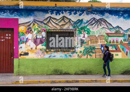 Une jeune fille marche à côté d'une peinture murale représentant la vie d'un village sur le côté d'une maison à San Antonino Castillo Velasco, Oaxaca, Mexique. La scène dep Banque D'Images