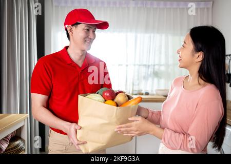 Homme de livraison asiatique en uniforme rouge donnant un sac en papier de nourriture, de fruits, de légumes à la cliente féminine à sa maison. Fournir un service d'épicerie à la maison Banque D'Images