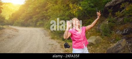 Bonne femme touriste senior marchant sur la route forestière d'été à l'extérieur à l'heure du coucher du soleil. Une vieille femme mince voyageant avec un appareil photo fait le selfie sur un smartphone. Concept de vacances de retraite actives. Filtre chaud Banque D'Images
