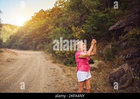 Bonne femme touriste senior marchant sur la route forestière d'été à l'extérieur à l'heure du coucher du soleil. Une vieille femme mince voyageant avec un appareil photo fait le selfie sur un smartphone. Concept de vacances de retraite actives. Filtre chaud Banque D'Images