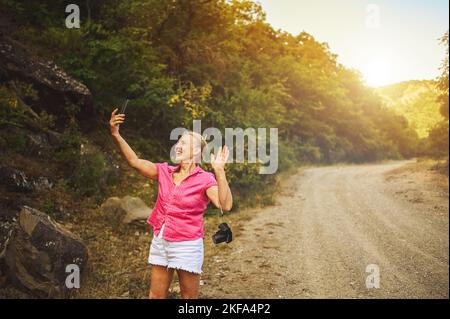 Bonne femme touriste senior marchant sur la route forestière d'été à l'extérieur à l'heure du coucher du soleil. Une vieille femme mince voyageant avec un appareil photo fait le selfie sur un smartphone. Concept de vacances de retraite actives. Filtre chaud Banque D'Images
